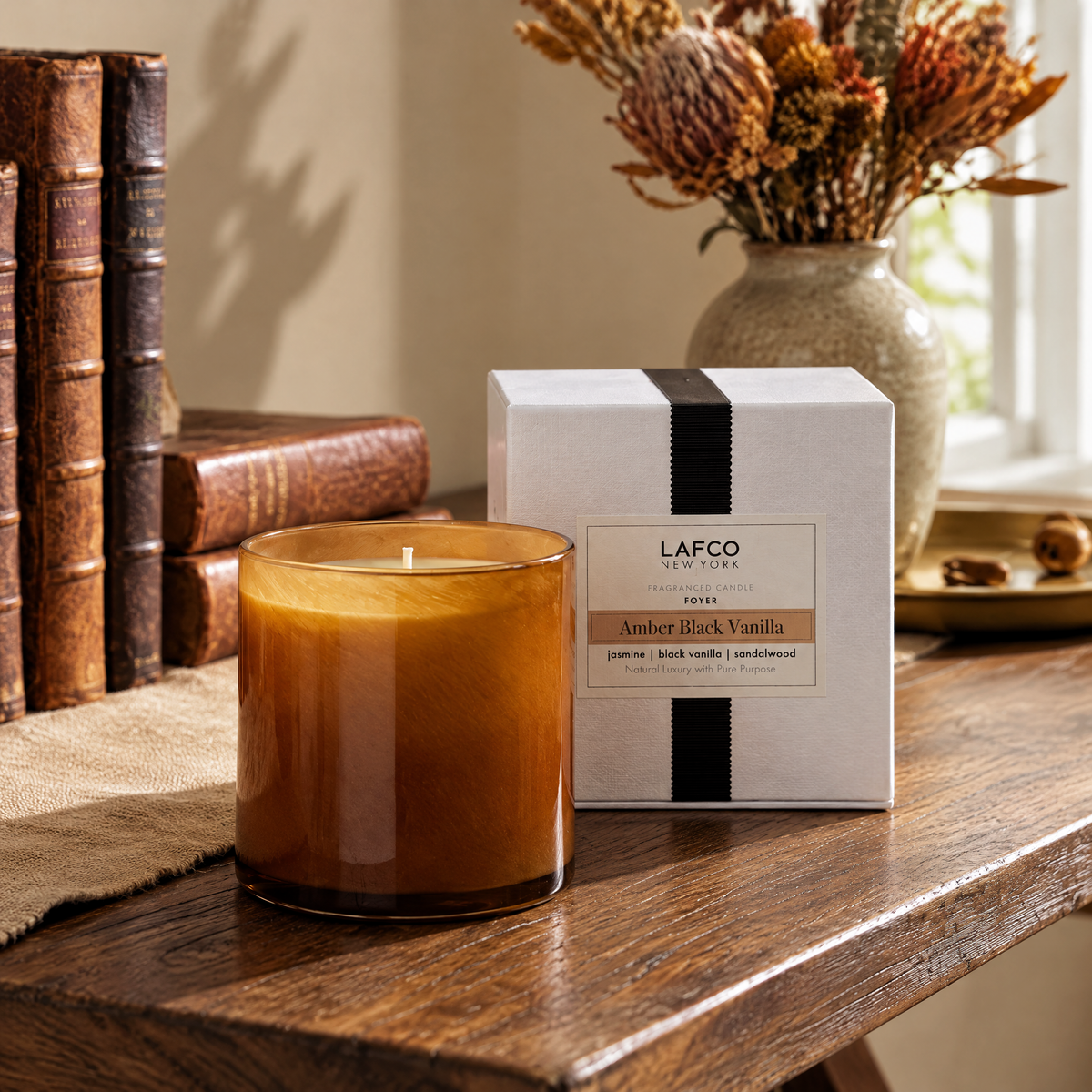 A LAFCO New York Amber Black Vanilla candle and box on a rustic wooden table with vintage books and dried flowers.