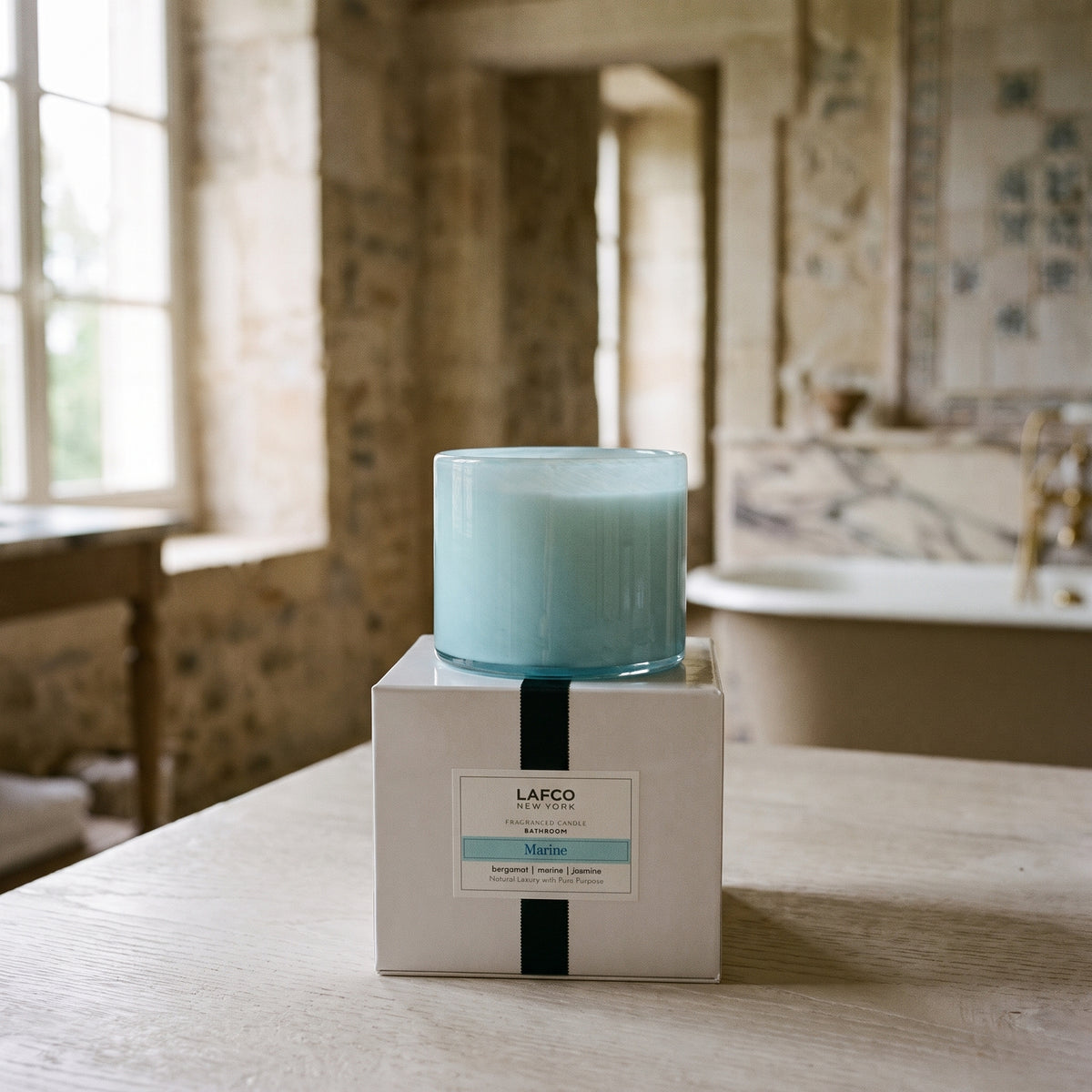 Lafco New York Marine candle on a white box in a rustic, sunlit stone bathroom with a vintage soaking tub.